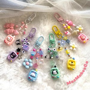 May include: A set of six colorful keychains featuring a variety of charms, including cartoon characters, flowers, stars, and beads. Each keychain has a small, rectangular charm with a cartoon character on it. The keychains are attached to silver key rings.