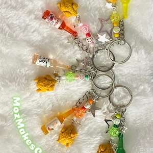 May include: A set of six colorful keychains featuring miniature bottles of Korean soju and fried chicken charms. Each keychain has a silver star charm and colorful beads.