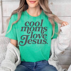 May include: A green t-shirt with the text "cool moms love Jesus" in dark gray lettering. The shirt is worn by a person with long blonde hair, wearing a gray cardigan and blue jeans.