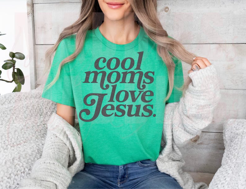 May include: A green t-shirt with the text "cool moms love Jesus" in dark gray lettering. The shirt is worn by a person with long blonde hair, wearing a gray cardigan and blue jeans.