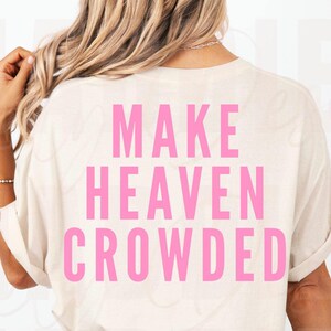 May include: A cream-coloured t-shirt with the words "MAKE HEAVEN CROWDED" printed in bright pink block letters. The shirt is viewed from the back, with rolled-up sleeves. The model is wearing a bracelet and has blonde hair.