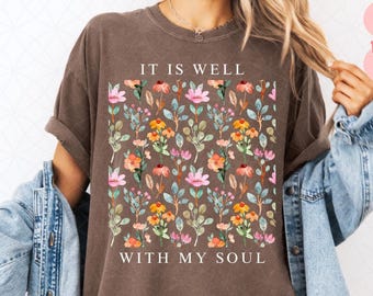 May include: A brown t-shirt with a floral pattern featuring pink, yellow, and orange flowers, and green leaves. The text "IT IS WELL WITH MY SOUL" is printed on the shirt in white.