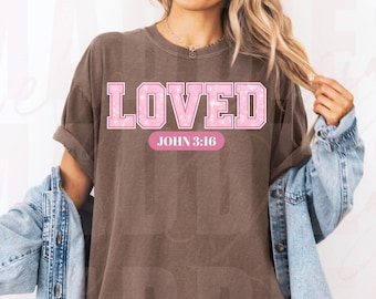 Puede incluir: Una camiseta marrón con la palabra "LOVED" en letras rosas con un estampado de estrellas blancas. Debajo de la palabra "LOVED" está el texto "JOHN 3:16" en un rectángulo rosa. La camiseta tiene las mangas enrolladas.