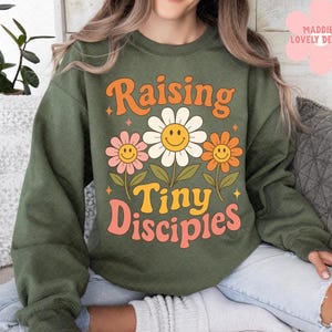 May include: Olive green sweatshirt with the text "Raising Tiny Disciples" in orange and pink. The design features three smiling daisy flowers in pink, white, and orange. The sweatshirt has a crew neck and long sleeves.
