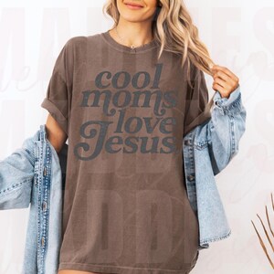 May include: Brown t-shirt with the text "cool moms love Jesus" in a dark gray font. The shirt is worn with a light blue denim jacket. The overall style is casual and comfortable.