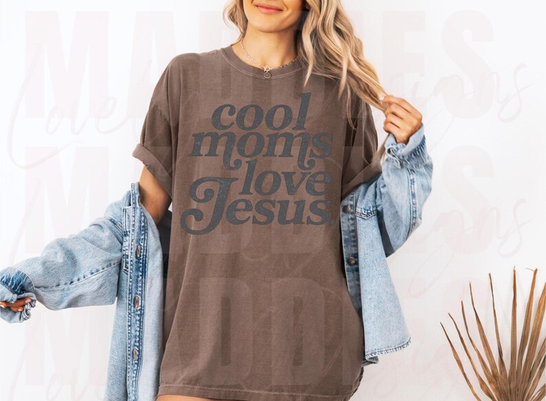May include: Brown t-shirt with the text "cool moms love Jesus" in a dark gray font. The shirt is worn with a light blue denim jacket. The overall style is casual and comfortable.