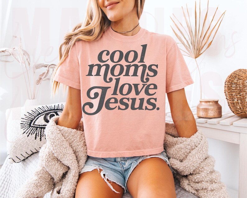 May include: A light pink t-shirt with the text "cool moms love Jesus" in a dark gray, stylized font. The shirt is paired with denim shorts and a cream-colored cardigan.