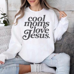 May include: White sweatshirt with the text "cool moms love Jesus" in dark gray lettering. The sweatshirt is a crew neck style and is being worn by a person. The person is wearing blue jeans.