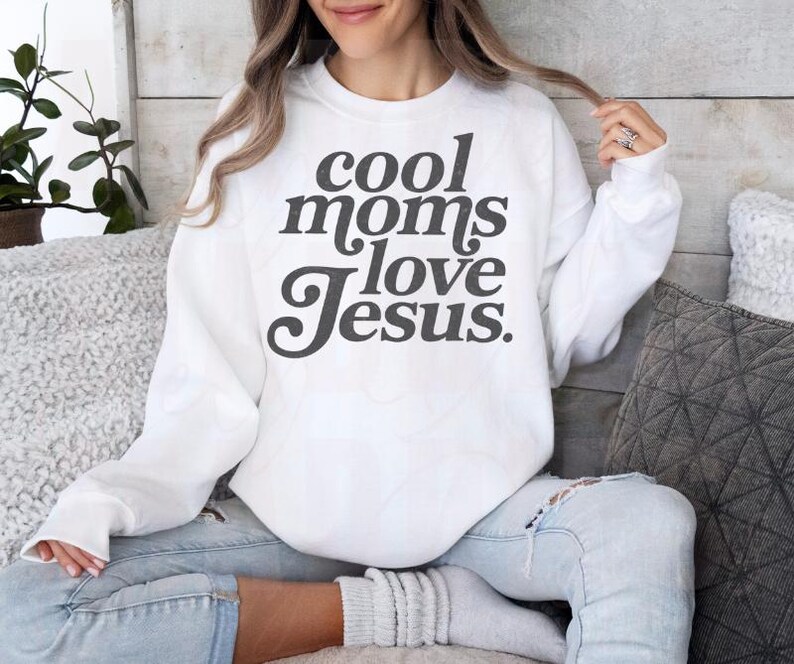 May include: White sweatshirt with the text "cool moms love Jesus" in dark gray lettering. The sweatshirt is a crew neck style and is being worn by a person. The person is wearing blue jeans.