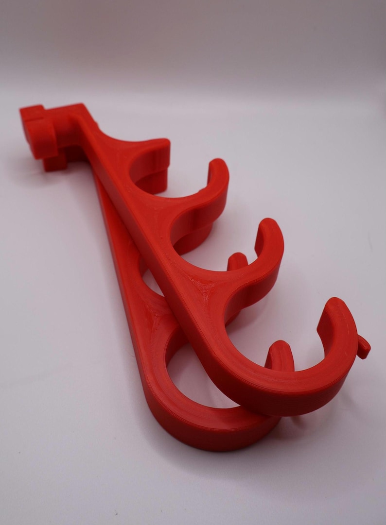 May include: A bright red plastic organizer rack featuring four curved hooks arranged in a vertical row. The structure has a mounting bracket at the top end and a series of circular cutouts along the main body, designed for hanging multiple items.