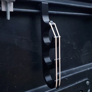 May include: A black, vertical, multi-hook storage accessory featuring three circular cutouts for organization. The hook is secured to a dark surface with a metal clamp and includes two light-colored elastic bands stretched across the front.
