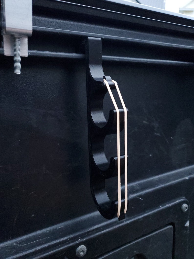 May include: A black, vertical, multi-hook storage accessory featuring three circular cutouts for organization. The hook is secured to a dark surface with a metal clamp and includes two light-colored elastic bands stretched across the front.