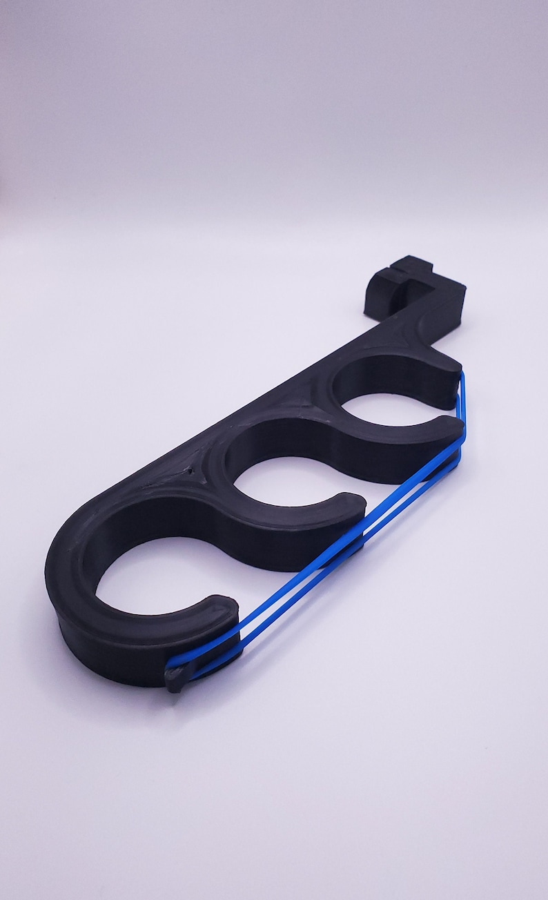 May include: A black, elongated, rigid plastic tool featuring three circular openings arranged in a row. A thin, bright blue elastic band is stretched across the front of the three circular openings, secured by small notches at each end of the frame.