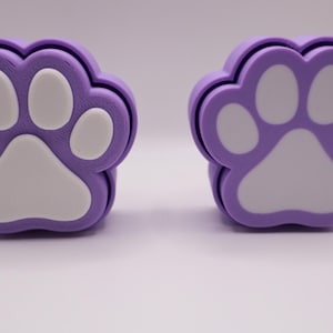 May include: Two purple paw-shaped coasters with white paw pad and toe details. The coasters are made of a soft material and are designed to protect surfaces from scratches and water rings. The paw print design is a popular theme for pet lovers.