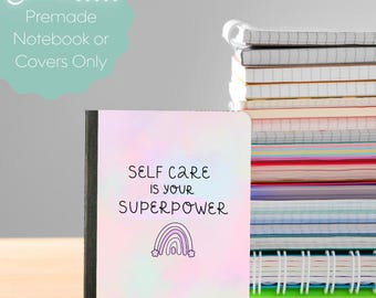 Può includere: Un taccuino con uno sfondo ad acquerello pastello che presenta il testo nero "SELF CARE IS YOUR SUPERPOWER" e una piccola illustrazione di un arcobaleno viola. Accanto c'è un'alta pila di vari taccuini a spirale con pagine colorate.