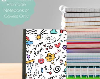 May include: A notebook with a white cover featuring colorful doodle art, including hearts, stars, and the word "love." The notebook is next to a stack of other notebooks. Text in the image reads "Choose... Premade Notebook or Covers Only."