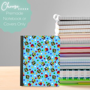 May include: A notebook with a blue cover featuring a pattern of colorful Rubik's Cubes. The text "Choose... Premade Notebook or Covers Only" is in the upper left corner. A stack of notebooks is visible in the background.