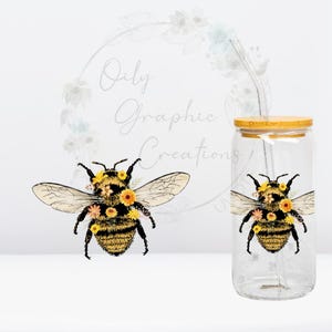 May include: A clear glass tumbler with a bamboo lid and a reusable straw. The tumbler features a detailed illustration of a bee adorned with flowers. The bee design is repeated on the tumbler. The text "Oily Graphic Creations" is in the background.