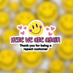 May include: A white sticker with a yellow smiley face and pink hearts. The text on the sticker reads "Here we are again. Thank you for being a repeat customer."