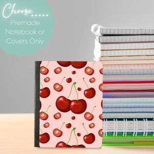 May include: A notebook with a cherry pattern on a light pink background. The cover features a repeating design of red cherries and cherry halves. The text "Choose... Premade Notebook or Covers Only" is in a light green bubble. A stack of notebooks is in the background.
