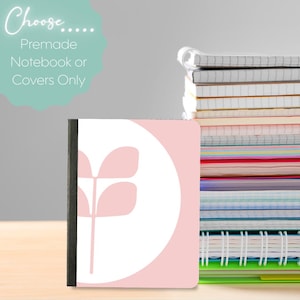 May include: A notebook with a pink cover featuring a white floral design and a black spine. The text "Choose... Premade Notebook or Covers Only" is in a teal bubble. A stack of notebooks is visible in the background.