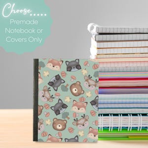 Puede incluir: Un cuaderno con un estampado de animales del bosque con zorros, mapaches, ciervos y osos sobre un fondo verde claro. El cuaderno forma parte de una pila de cuadernos y tiene el texto "Choose... Premade Notebook or Covers Only".