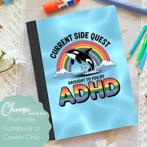 May include: A notebook with a light blue cover featuring a cartoon orca leaping through a rainbow. The text reads "CURRENT SIDE QUEST BROUGHT TO YOU BY ADHD" in black and rainbow colors. The notebook is black and has the text "Choose Premade Notebook or Covers Only".