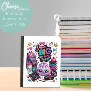 May include: A white notebook cover featuring a colorful graphic with the text Dream Big, Work Hard, Stay Kind. The illustration includes a backpack, a cup, a stack of books, a pencil case, headphones, and a smiling purple dumpling character.