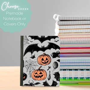 May include: A Halloween-themed notebook with a gray cover featuring black bats, spiderwebs, and jack-o'-lanterns. The text "Choose... Premade Notebook or Covers Only" is displayed above the notebook. A stack of notebooks is visible in the background.
