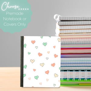 Puede incluir: Una cubierta de cuaderno blanca con un patrón repetitivo de pequeños corazones dibujados a mano en tonos pastel melocotón, verde menta y amarillo. El texto dice: Choose..... Premade Notebook or Covers Only. Se ve una pila de cuadernos.
