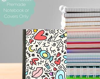 May include: A colorful notebook with a whimsical doodle design featuring hearts, stars, and candy on a cream background. The notebook is next to a stack of other notebooks. The text in the image says "Choose... Premade Notebook or Covers Only."