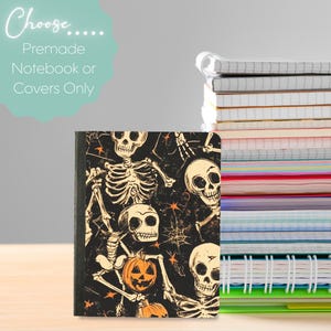 May include: A notebook with a black cover featuring a repeating pattern of white skeletons, orange jack-o'-lanterns, and small orange stars. To the right, a stack of various notebooks with white spiral bindings and colorful pages is visible.