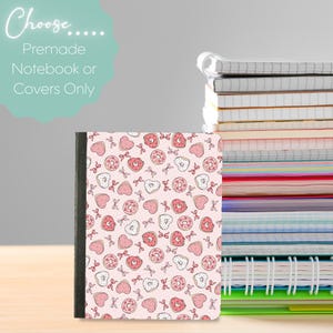 May include: A notebook with a pink cover patterned with donuts, hearts, and bows. The notebook has a black spine and is next to a stack of other notebooks. The image includes the text "Choose... Premade Notebook or Covers Only."