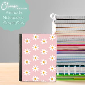 Könnte beinhalten: Ein rosa Notizbuchumschlag mit einem Gänseblümchenmuster, das weiße Blüten mit gelben Zentren zeigt. Der Text "Choose... Premade Notebook or Covers Only" befindet sich oben links. Ein Stapel Notizbücher ist im Hintergrund zu sehen.