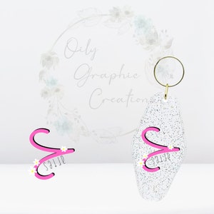 May include: A white glitter keychain with a pink letter "S" and white flowers. The keychain is in the shape of a motel key. The text "Oily Graphic Creations" is visible in the background.