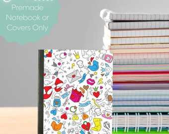 May include: A notebook with a white cover featuring a colorful doodle design of hearts, bows, birds, and other whimsical elements. The notebook has a black spine and is part of a stack of notebooks. Text on the cover reads "Choose... Premade Notebook or Covers Only."