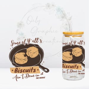 May include: A clear glass jar with a bamboo lid and a straw. The jar has a graphic of a cast iron skillet with biscuits inside. The text on the jar reads "Some of Y'all's Biscuits. Ain't Done in the Middle."
