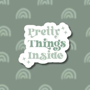 May include: A white sticker with the text "Pretty Things Inside" in a light green font. The sticker has a cloud-like shape and is decorated with small star shapes. The background is a muted green color with a pattern of blurred rainbow shapes.