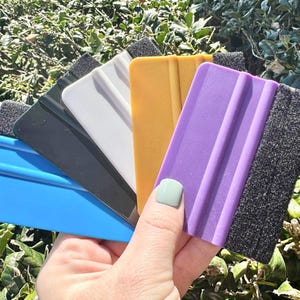 Puede incluir: Una colección de rasquetas de colores en azul, negro, blanco, amarillo y morado. Cada rasqueta tiene forma rectangular con un borde de espuma. Las herramientas se sostienen sobre un fondo de follaje verde.