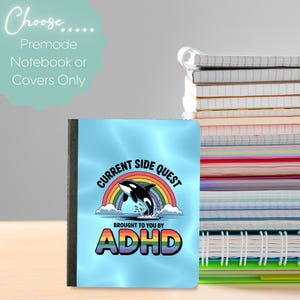 Peut inclure: Un cahier avec une couverture bleue représentant un arc-en-ciel, une orque sautant et le texte "CURRENT SIDE QUEST BROUGHT TO YOU BY ADHD". Le cahier est à côté d'une pile d'autres cahiers.