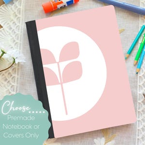 May include: A pink notebook with a white circle and a light pink floral design. The notebook has a black spine and the text "Choose... Premade Notebook or Covers Only". Colored pencils and glue stick are visible.
