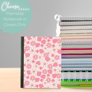 May include: A notebook with a pink cover featuring a pattern of strawberries, daisies, and butterflies. The notebook has a black spine and is part of a stack of notebooks. Text in the image reads "Choose... Premade Notebook or Covers Only."