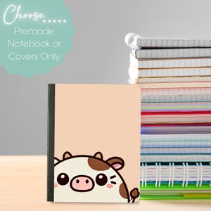 Puede incluir: Un cuaderno con una cubierta melocotón claro con un diseño de vaca de dibujos animados. El cuaderno tiene un lomo negro y está junto a una pila de cuadernos coloridos. El texto a la izquierda dice "Choose... Premade Notebook or Covers Only."