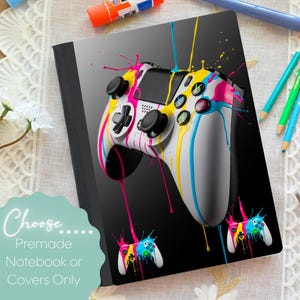 Puede incluir: Cubierta de cuaderno con un diseño de mando de videojuego blanco y salpicaduras de pintura coloridas en amarillo, rosa y azul. La cubierta tiene el texto "Choose... Premade Notebook or Covers Only".