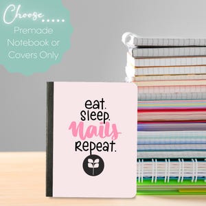 May include: A notebook with a light pink cover featuring the text "eat. sleep. nails. repeat." in black and pink. A stack of notebooks is visible in the background. The notebook has a black spine and a black and white logo at the bottom.