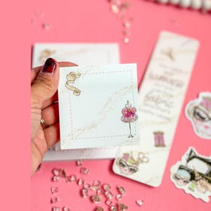 May include: A square white sticky note featuring a dashed border, a gold ribbon illustration in the top left corner, and a pink dress on a mannequin stand in the bottom right corner, accented by a subtle gold glitter swirl design across the paper.