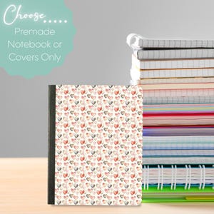 Puede incluir: Un cuaderno con una cubierta blanca que presenta un patrón repetitivo de pequeños corazones de acuarela en tonos rosa, rojo y gris, colocado junto a una alta pila de varios cuadernos de espiral con páginas y cubiertas coloridas.