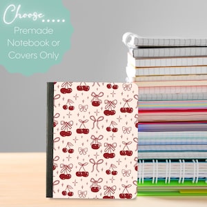 Op de afbeelding: Een notitieboek met een crèmekleurige kaft met een patroon van rode kersen en strikken. Het notitieboek heeft een zwarte rug en maakt deel uit van een stapel notitieboeken. De tekst "Choose... Premade Notebook or Covers Only" staat linksboven.
