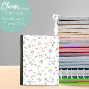 Puede incluir: Un cuaderno con cubierta blanca que presenta un patrón repetitivo de patos de dibujos animados con botas de lluvia azul claro y pequeñas flores rosas. El texto dice Choose..... Premade Notebook or Covers Only. Se ve una pila de cuadernos.