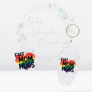 Könnte beinhalten: Ein Paar glitzernde, transparente Acryl-Ohrringe mit einem goldfarbenen Ring. Jeder Ohrring hat ein Regenbogenherz mit den schwarzen Worten "FREE MOM HUGS". Ein einzigartiges Accessoire.